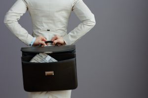 Seen from behind female worker in a light business suit with briefcase and dollars money packs isolated on grey.